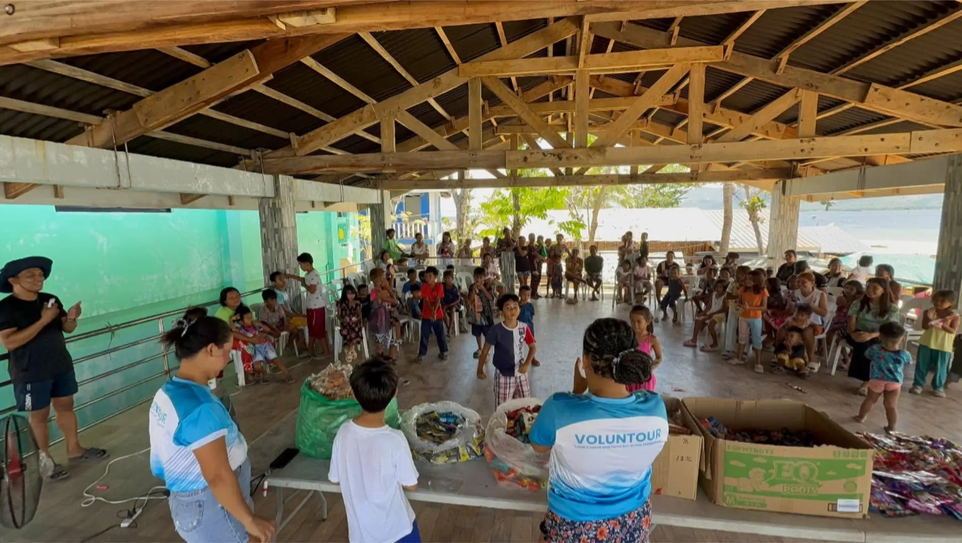 Volunteers in blue shirts interacting with children and families inside a covered wooden structure on Mararisaon Island.