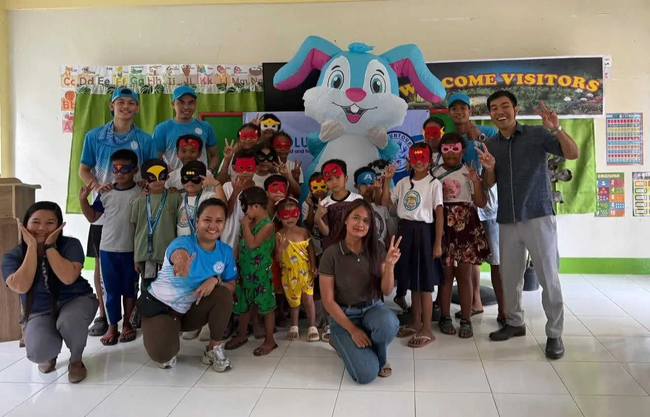 Smaller group of children and volunteers posing indoors with a large cartoon mascot figure and bright decorations behind them.