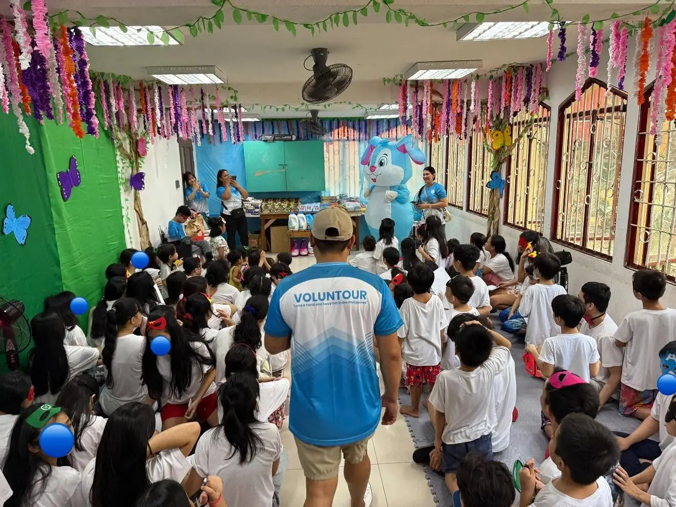Classroom filled with children seated and facing the front while a volunteer in a blue and white shirt leads an activity or presentation.