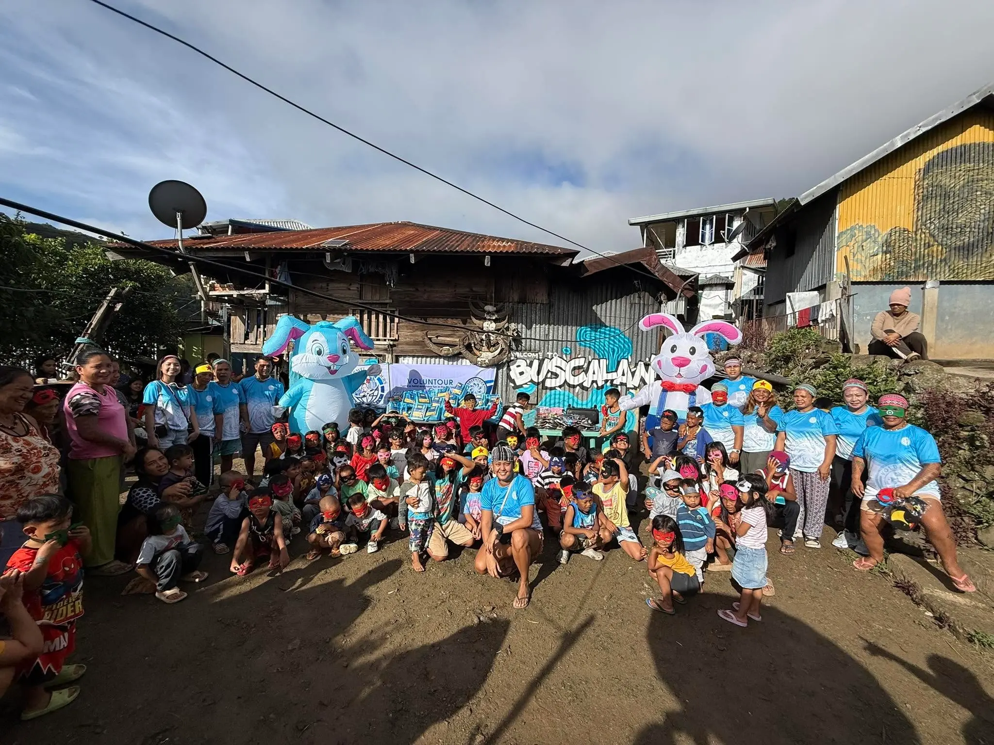 Large group of volunteers in blue shirts gathered outdoors in front of a decorated wall with balloons during a community event.