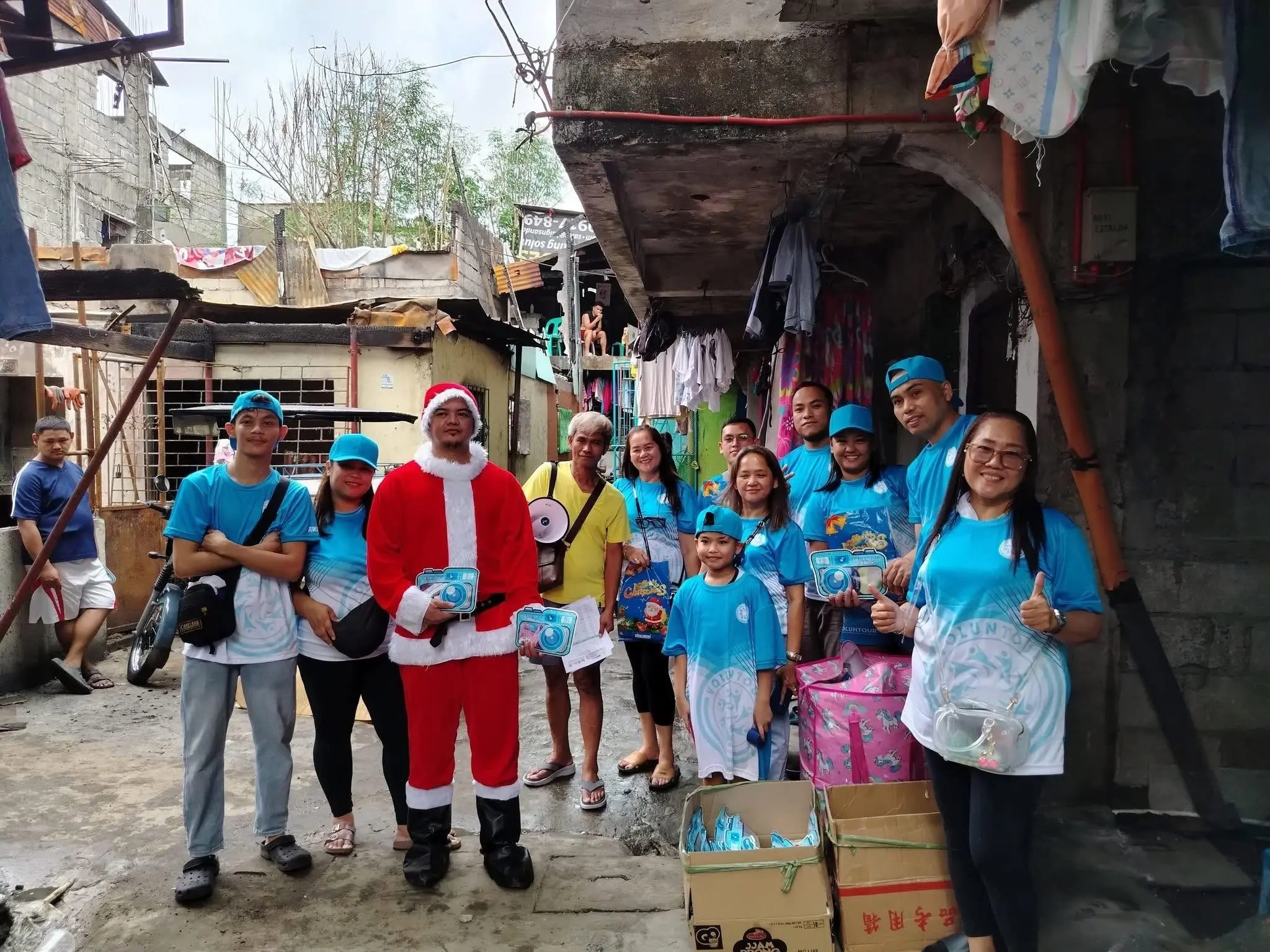 Volunteers in blue shirts standing in a narrow street with one dressed as Santa Claus and others holding boxes for a festive outreach activity.