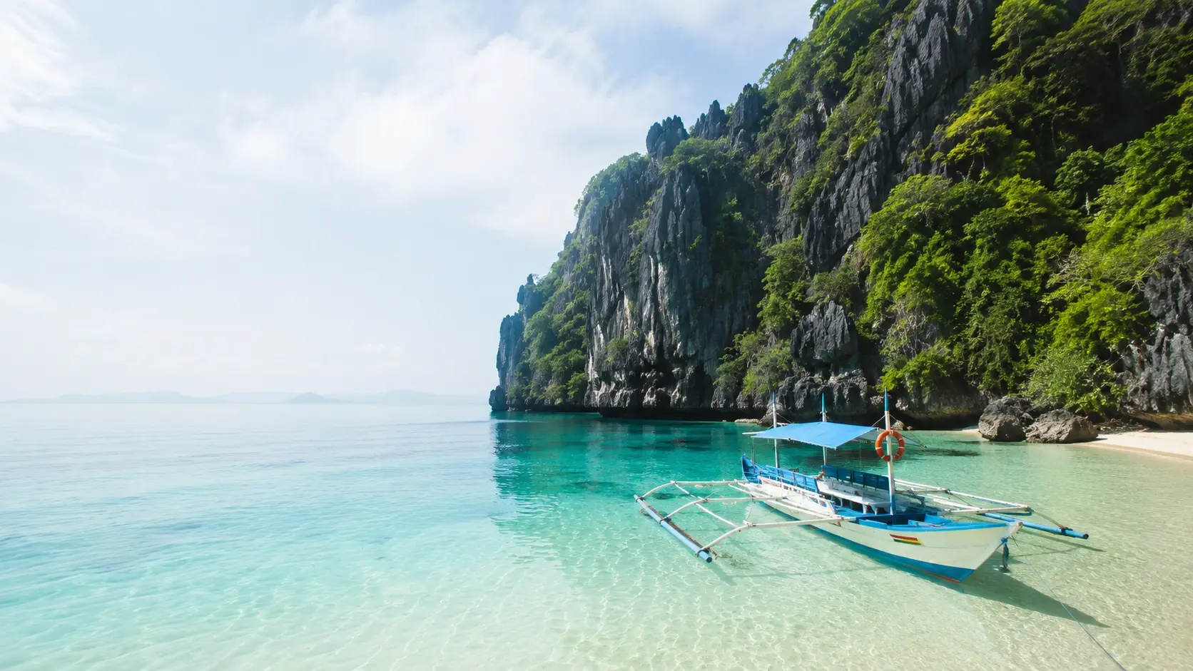 Traditional outrigger boat floating in clear turquoise water near a sandy beach with limestone cliffs and lush greenery, symbolizing cultural heritage and natural beauty in the Philippines.
