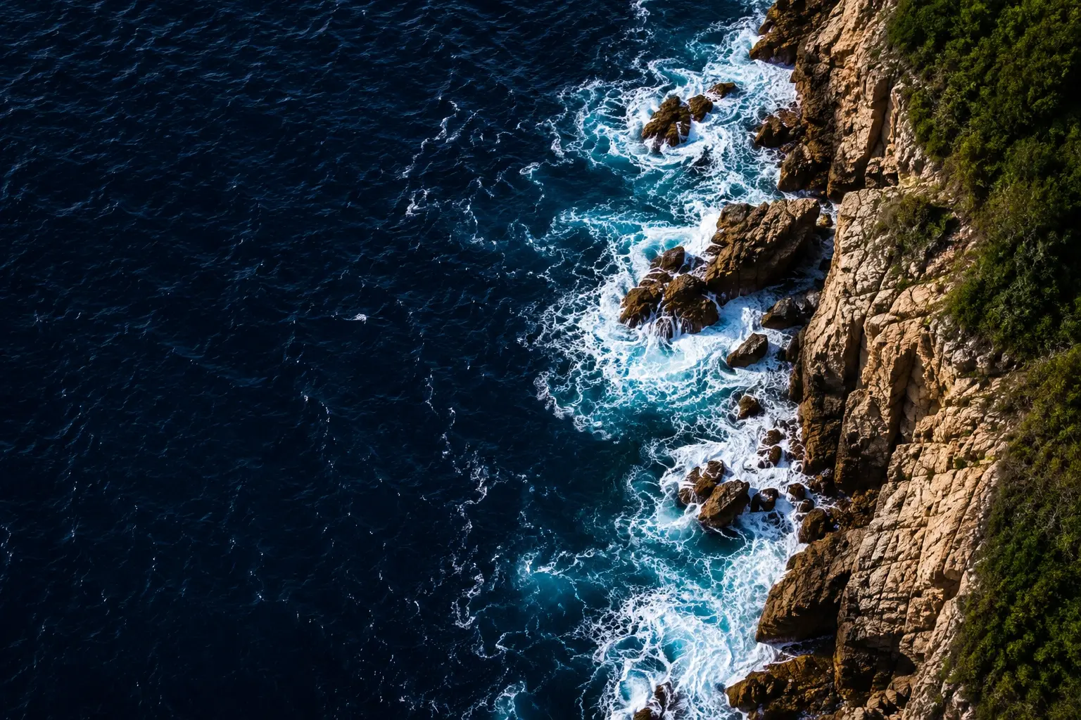 Aerial view of deep blue ocean waves crashing against rocky cliffs with white foam and green vegetation, symbolizing nature’s power and conservation for Voluntour Philippines.