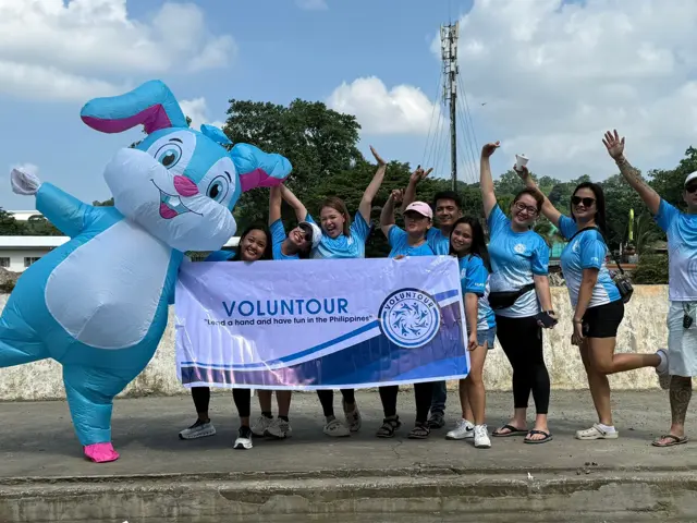 Volunteers working together outdoors in the Philippines, holding a Voluntour banner with mascot character.